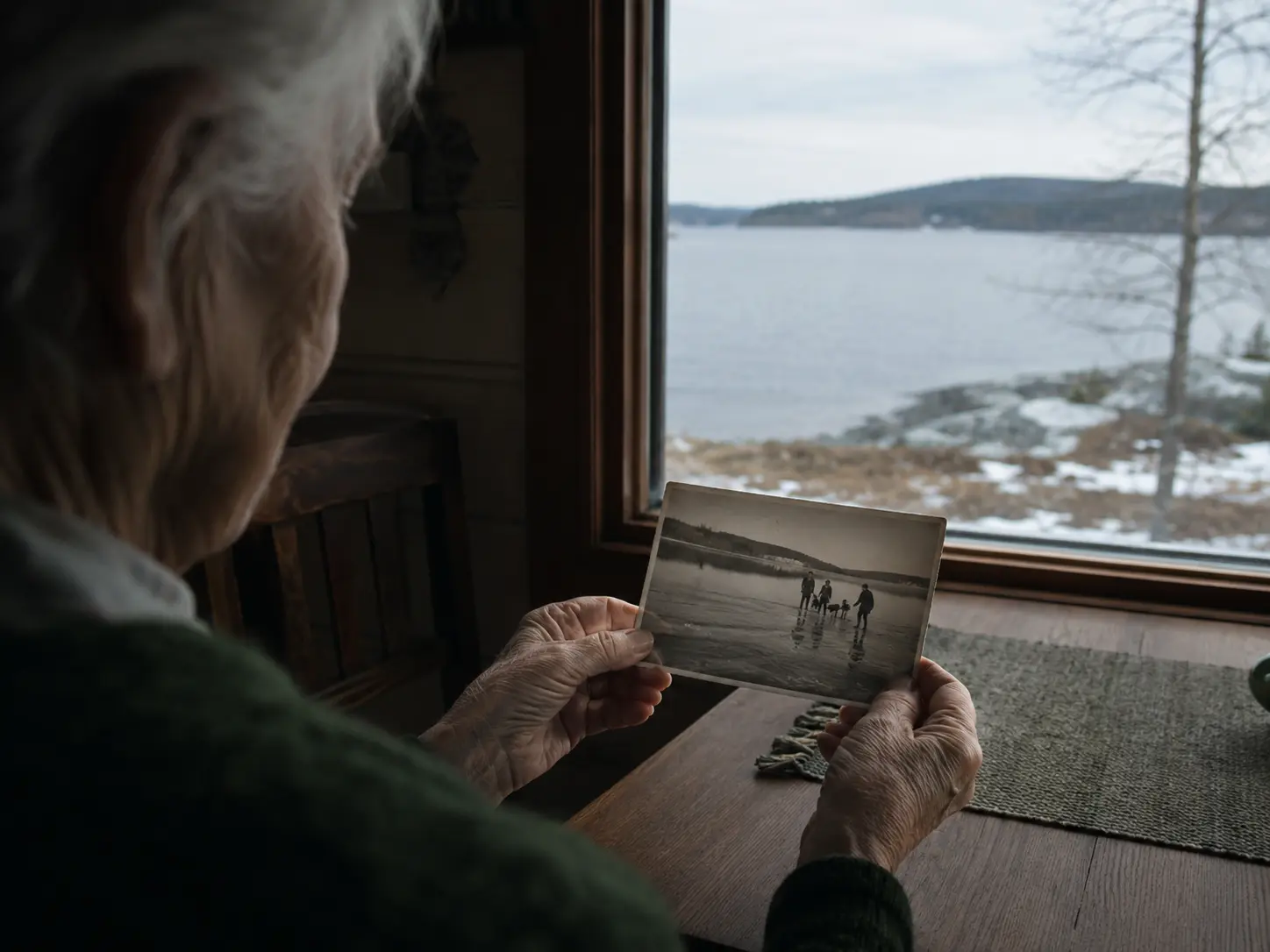 An older person holding an old winter photograph beside a present-day northern lake